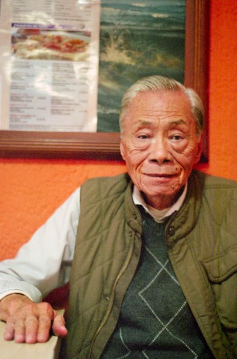 An elderly Chinese man beams while sitting at a table in his restaurant.