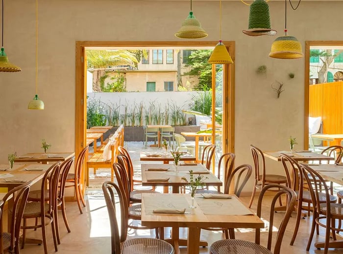 A bright dining room featuring large doors that open onto a patio, with wooden tables set for dinner and cozy pillows lining a bench on one side.