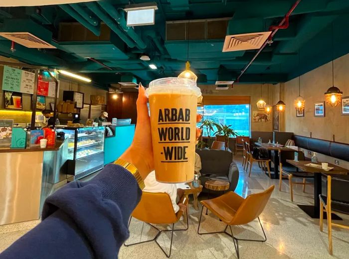 A hand lifts a plastic coffee cup featuring the branding 'Arab World Wide.'