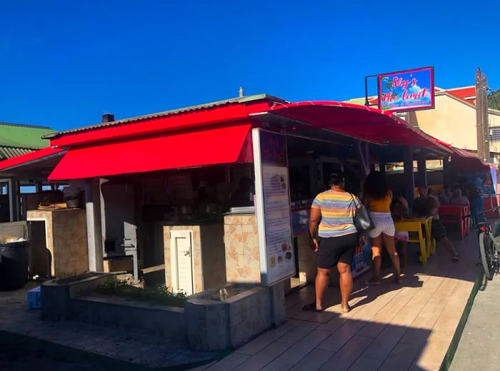 Patrons wait in line outside a beachside shack restaurant topped with a large red awning.