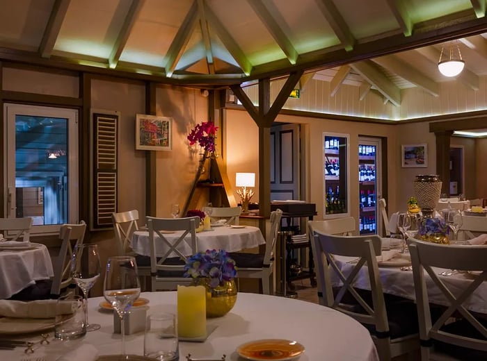 The interior of a restaurant adorned with white tablecloths, featuring high ceilings with wooden beams and lush greenery visible through the windows.