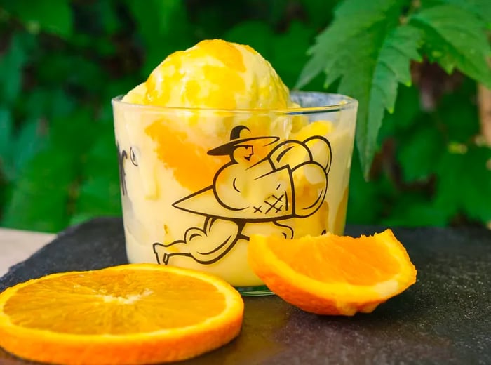 Scoops of orange sorbet served in a glass adorned with an illustration of a man in a wide-brimmed hat running with a giant sorbet cone, accompanied by orange slices.