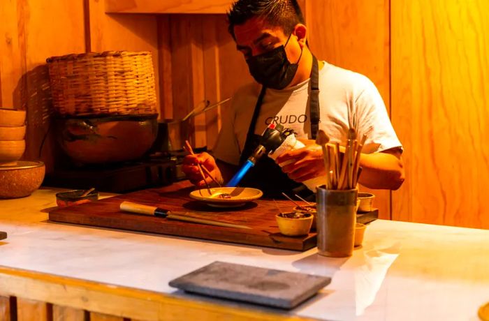 A masked chef employs a blow torch to prepare an ingredient.