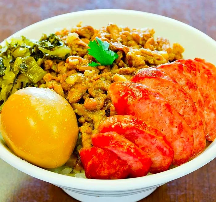 A bowl of rice topped with bright red slices of sausage, minced pork, a vividly colored egg, and stewed greens