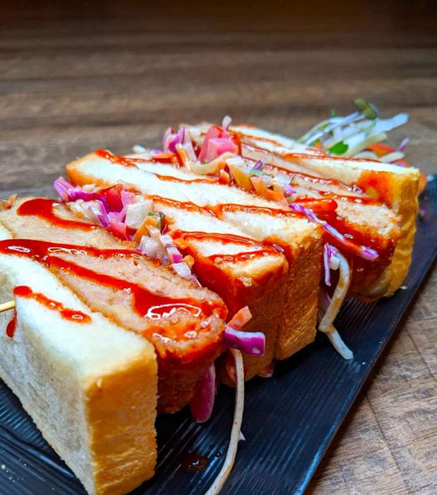 A crispy fried chicken sandwich served on thick bread, cut in half and presented on its side, topped with a drizzle of red sauce