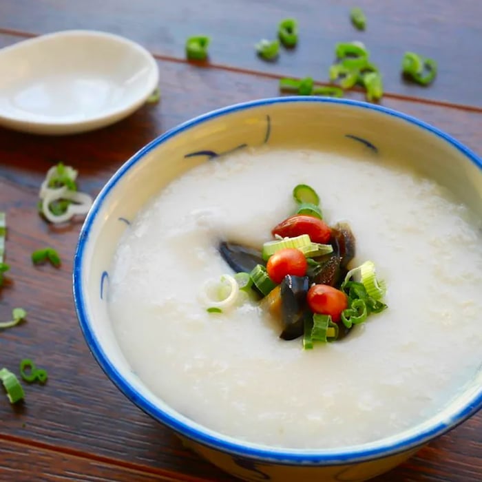 A bowl of congee showcasing a small heap of toppings peeking out from the center, placed on a wooden table sprinkled with chopped herbs