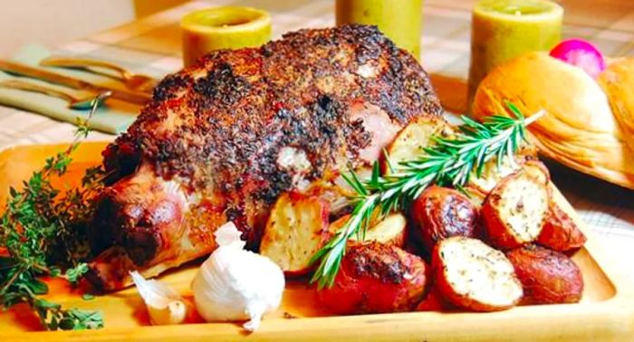 A substantial roast displayed on a cutting board, accompanied by sliced potatoes and fresh herbs.