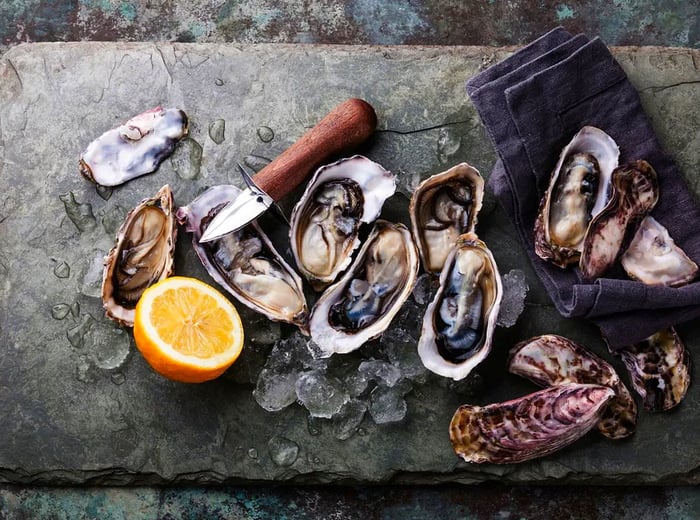 A top-down view of open and closed oysters on a slate cutting board, accompanied by an oyster knife and half a lemon