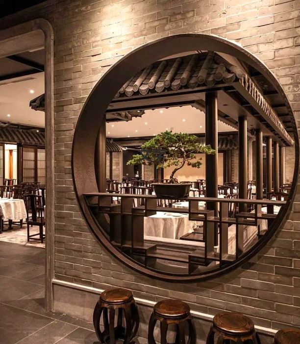 A restaurant interior featuring a large circular cutout in a brick wall, revealing a dining area that centers around an impressive bonsai tree