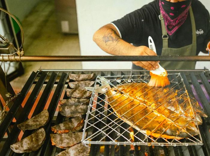 A chef applies sauce to a fish held on a grill by a metal grate.