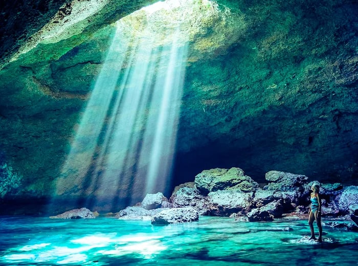 The author stands inside a massive cave, perched on a rock just above the water, gazing up at radiant beams of light streaming through an opening above. The light dances on the water's surface, creating a mesmerizing turquoise glow, with the entire scene enveloped in a greenish hue.