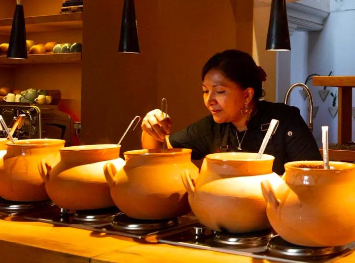 A chef carefully stirs a clay pot, one among several lined up beneath warm pendant lights on the counter.