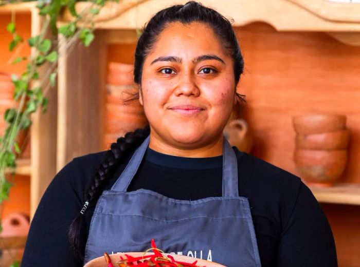 A chef in a branded apron holds a bowl of chiles, standing in front of shelves lined with clay pots.