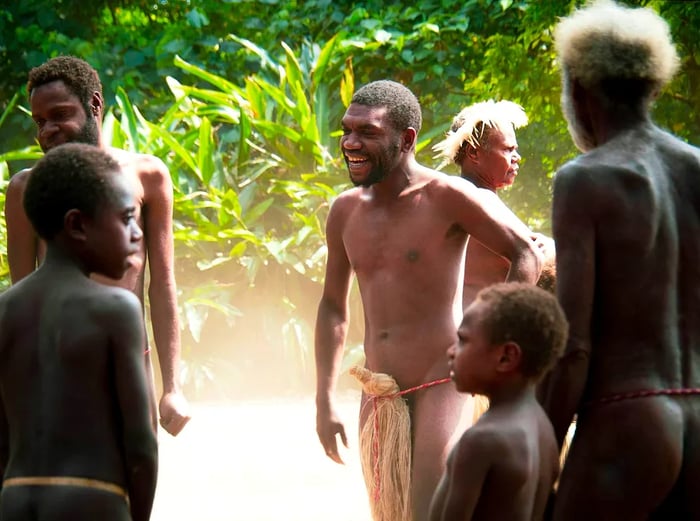 A group of men from the village of Yakel stands in a circle, dressed only in a string around their waists with a bundle of dried grass covering their groins; lush vegetation surrounds them, and dust swirls around the ground, likely kicked up from their dancing.