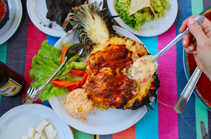 A spoon lifts gooey cheese from the center of a halved roasted pineapple.