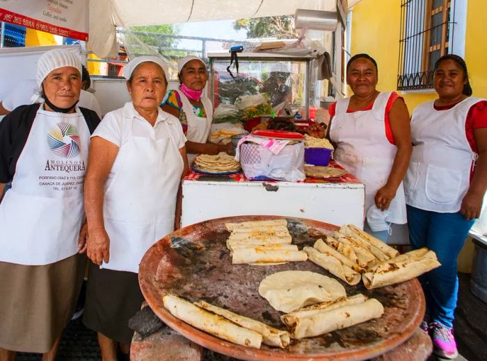 A group of women in aprons gather around a comal filled with numerous tacos.