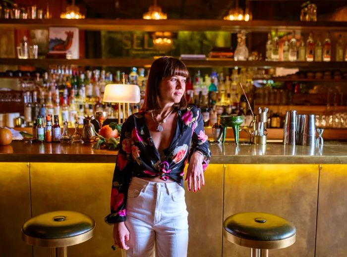 A woman dressed in a blouse and white pants stands in front of the bar.