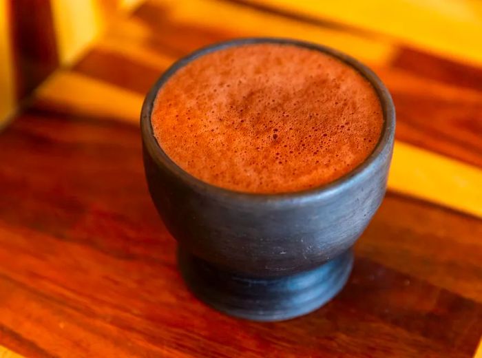 A frothy cup of drinkable chocolate in a black clay mug resting on a wooden table.