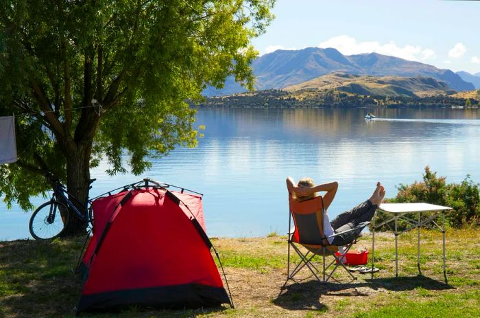 A woman enjoys relaxation at a lakeside campsite, next to her tent and bike, in New Zealand.