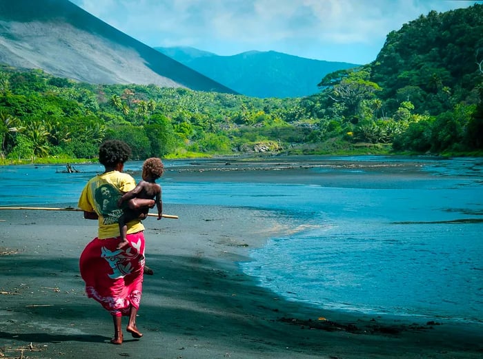 A local woman in a red sarong and a yellow Bob Marley t-shirt carries a child as she strolls along the riverbank of black sand; in the backdrop looms Mt Yasur with a tropical jungle at its base.