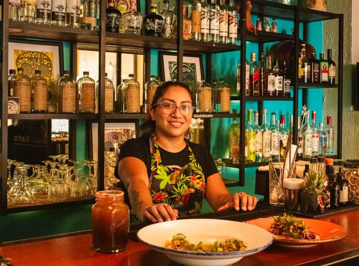 A woman beams from behind a bar where two dishes are presented.