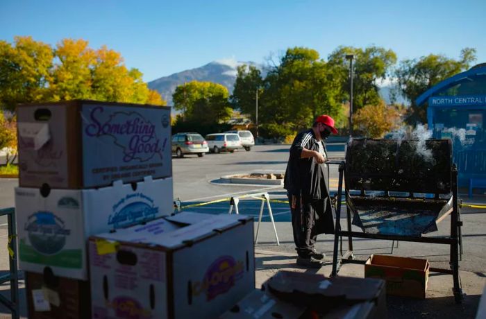 A man wearing a red cap and mask tends to a smoking metal chile roaster in a parking lot, with boxes of chiles nearby.