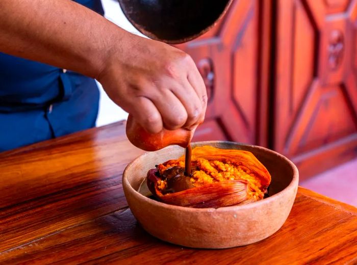 A chef lifts the lid from an earthenware pot, drizzling a rich dark sauce over a mashed dish wrapped in a corn husk.