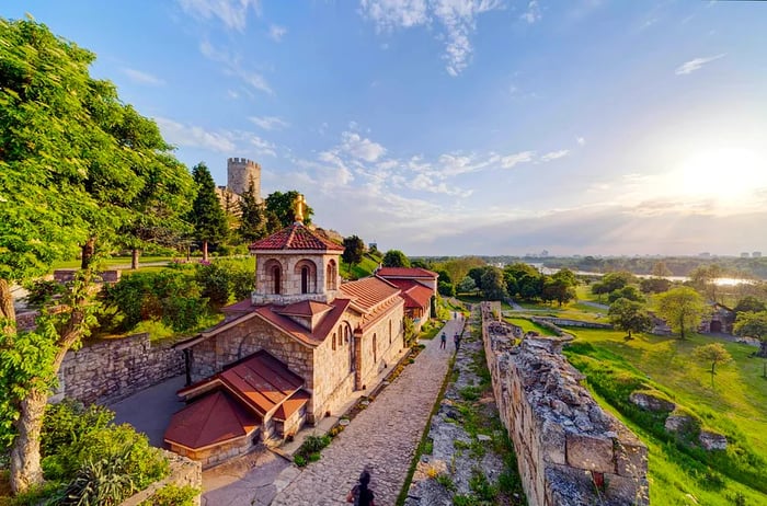 A grand fortress by the river with visitors strolling through the surrounding parkland.