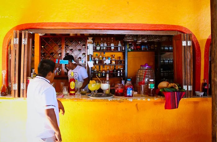 A waiter stands at the entrance of a yellow bar as a man places cocktails on a tray.