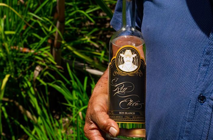 A brown hand grasps a bottle of clear rum labeled Rey Eteco.