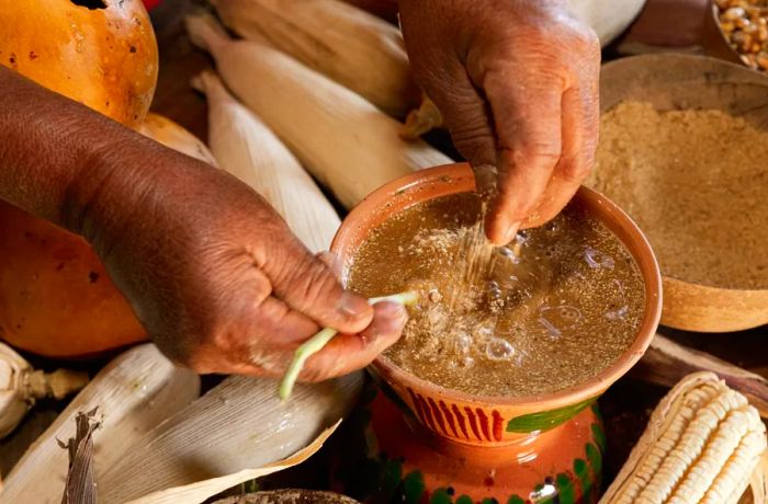 Worn hands blend roasted ground corn into a bowl of brown liquid.