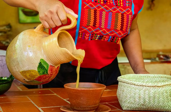 A woman in a traditional woven shirt pours a golden liquid from a ceramic pitcher into a sturdy ceramic bowl.