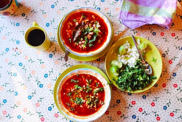 Two bowls of vibrant red pancita de res rest on a floral tablecloth.