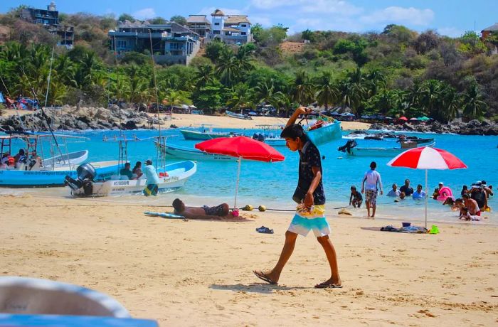 A boy carries a basket of food on a beach filled with umbrellas, people splashing in the water, and boats drifting by.