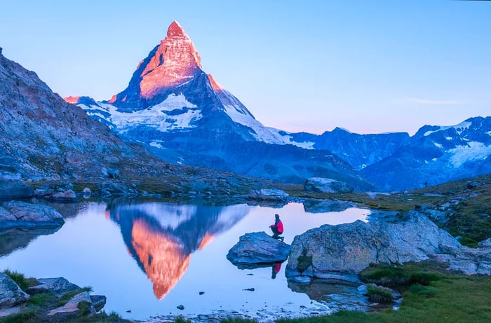 A breathtaking early morning scene of the Matterhorn at sunrise, casting a pink reflection on the lake, featuring a man in a red down jacket on a rock under a clear blue sky, Zermatt