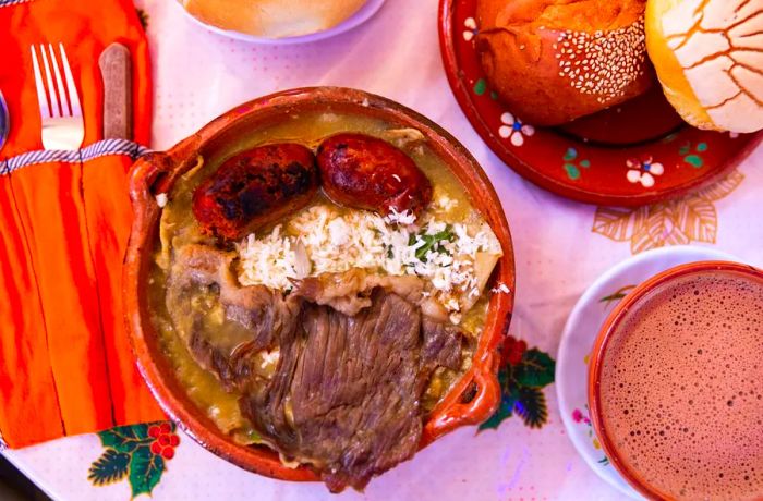 a dish served in a clay pot topped with meat, paired with a mug of hot chocolate and some pastries