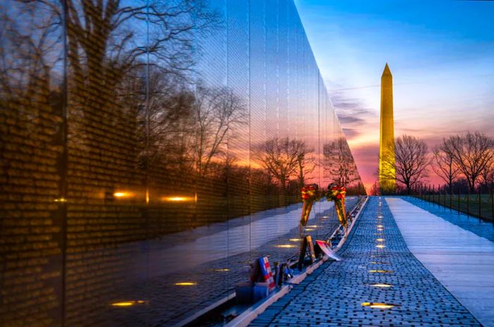 The early morning light shines on 'The Wall,' a prominent memorial inscribed with the names of those who perished in the Vietnam War, with a tall stone obelisk in the background.