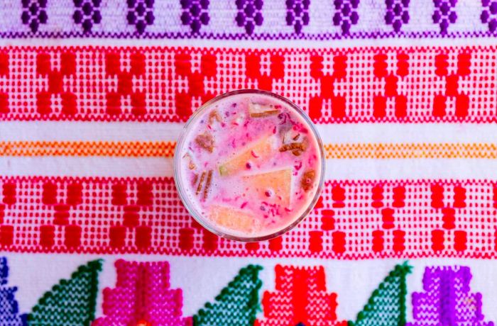 An aerial view of a cup filled with a pink creamy liquid adorned with nuts and melon, set on a vibrant blanket.