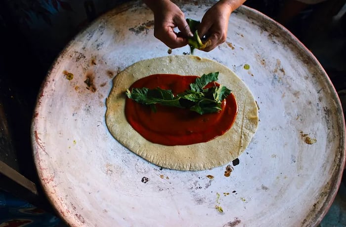 A large disc of masa topped with red sauce and herbs is sizzling away on a comal.