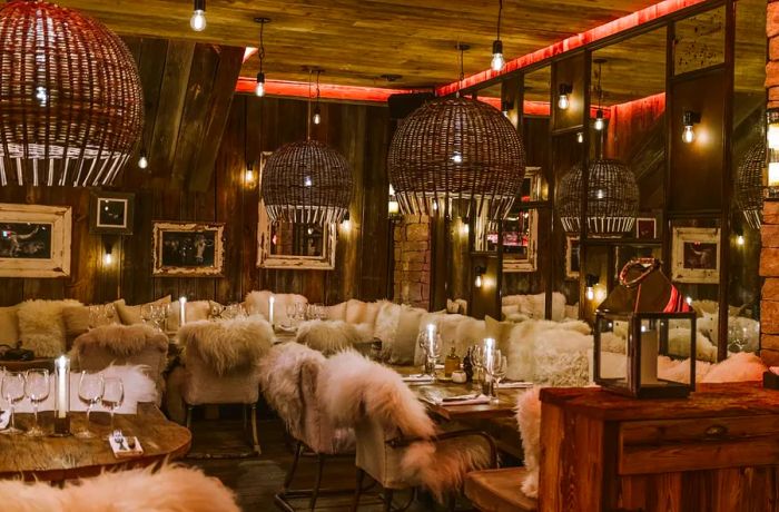 The restaurant boasts a cozy interior adorned with fur-covered chairs, intricate wooden pendant lights, candles flickering on dining tables, and warm wood paneling lining the walls.