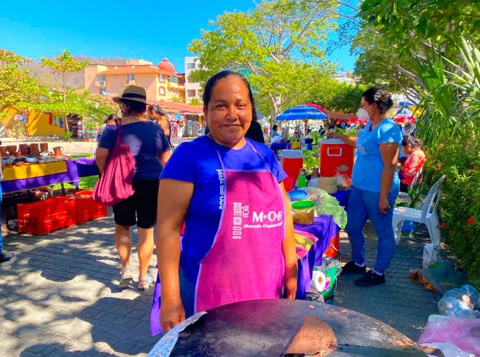 A woman in an apron stands by a comal in a park setting.