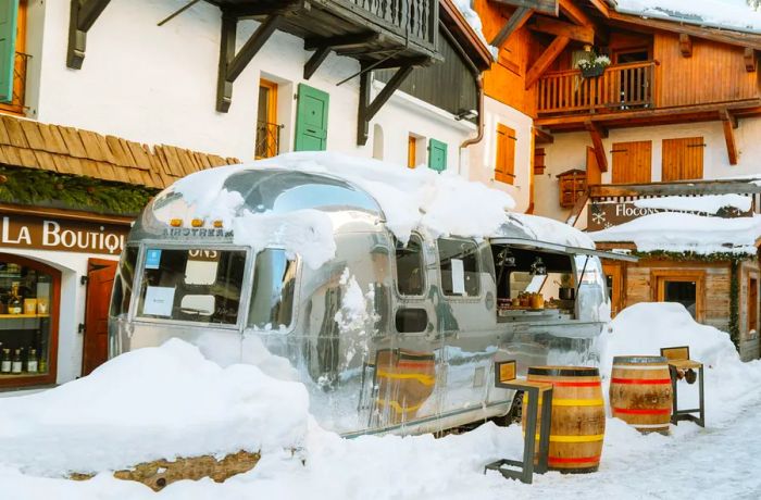 An Airstream trailer, blanketed in snow, parked outside a restaurant, accompanied by barrels set up as tables in a cozy dining area beside the trailer.