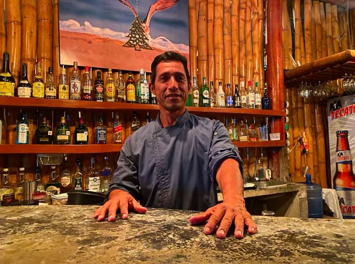 A man stands with his hands resting on a marble countertop, looking towards the camera, surrounded by shelves filled with bottles.