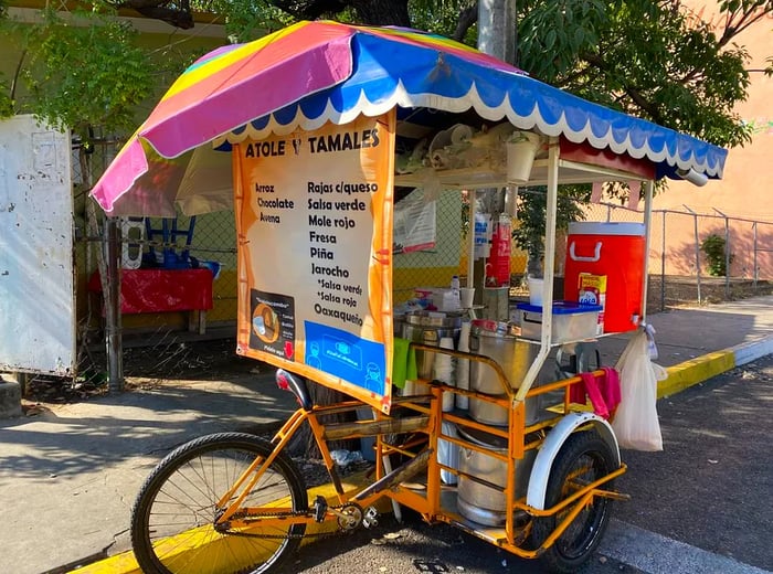 A tamale cart mounted on a bike, featuring a large, vibrant menu in front, a rainbow umbrella for shade, a cooler for drinks, and various kitchen tools at the ready.