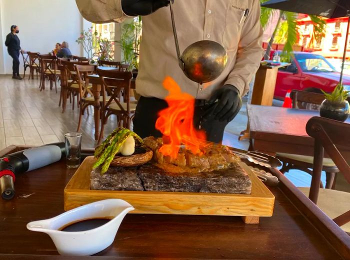 A server ignites sauce over a steak presented on a rustic wooden board.