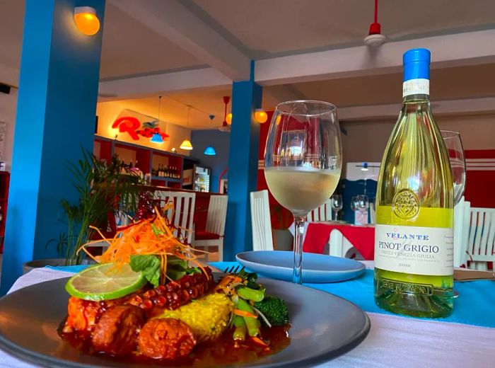 A dish of octopus surrounded by various garnishes in a rich sauce, accompanied by a glass and bottle of white wine, set in a stylish restaurant ambiance.