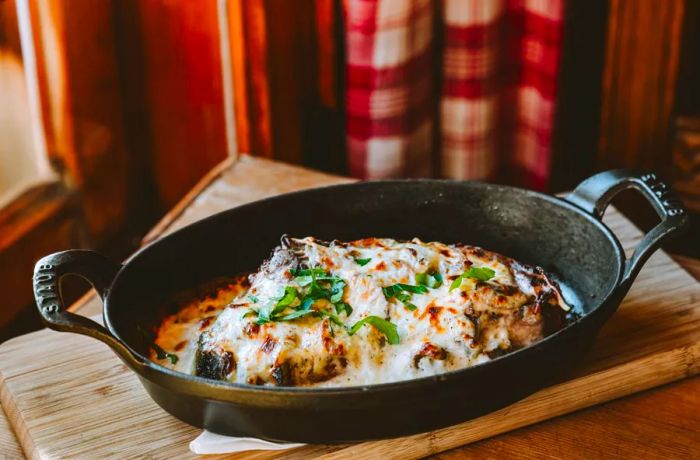 A cast iron pan brimming with cheese-topped bread and mushrooms, garnished with chopped herbs, rests on a wooden table.