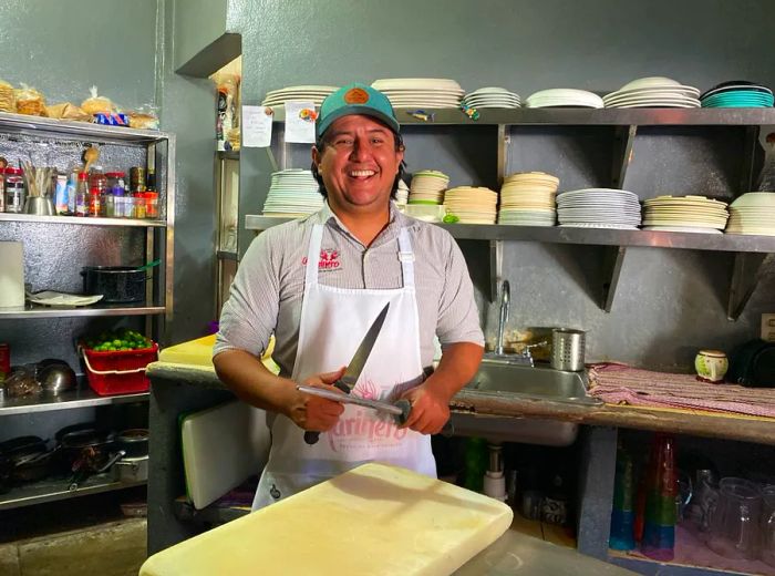 A chef holds knives in a kitchen, surrounded by plates and ingredients on shelves behind him.