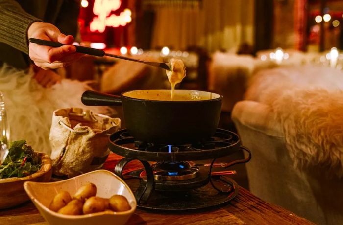 A hand uses a long skewer to dip a potato into a bubbling pot of fondue.