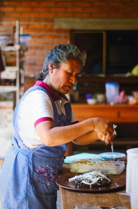 A woman in an apron adds a sprinkle of shredded cheese atop a dish of mole negro.